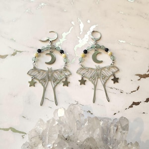 May include: A pair of silver-toned earrings featuring a moth design with dangling stars. Each earring has a crescent moon and is adorned with beads in shades of green, black, and yellow. The earrings are displayed on a white surface.