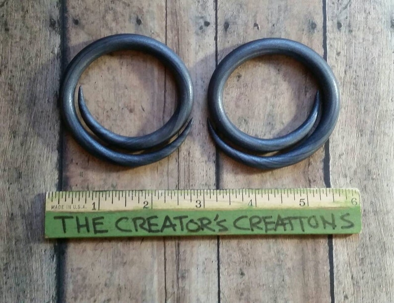 Peut inclure: Deux boucles d'oreilles en m&eacute;tal noir avec un design torsad&eacute;. Les boucles d'oreilles sont pr&eacute;sent&eacute;es sur une surface en bois avec une r&egrave;gle qui indique "The Creator's Creations".