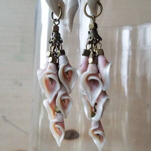 May include: A pair of white and pink flower-shaped dangle earrings with gold accents. The earrings are made of polymer clay and are attached to a pair of white plugs.
