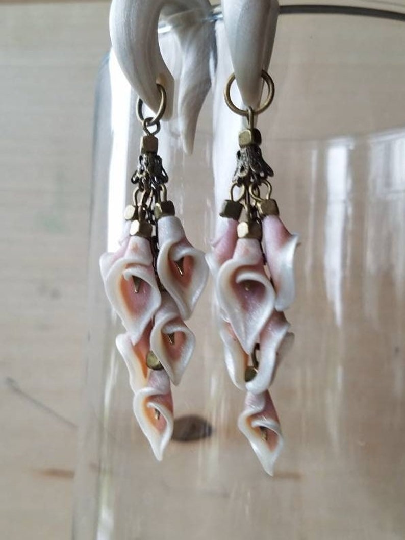 May include: A pair of white and pink flower-shaped dangle earrings with gold accents. The earrings are made of polymer clay and are attached to a pair of white plugs.