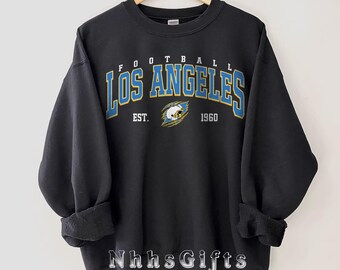 May include: Black sweatshirt with the text "FOOTBALL LOS ANGELES" in blue and gold lettering. Below the text is "EST. 1960" and a graphic of a football helmet. The sweatshirt is hanging on a wooden hanger.