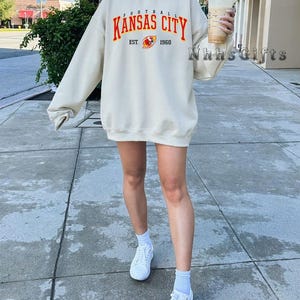 May include: Cream-colored sweatshirt with the text "KANSAS CITY FOOTBALL" in red and orange, with "EST. 1960" below. The sweatshirt is worn with white sneakers and socks. The image is taken outdoors.