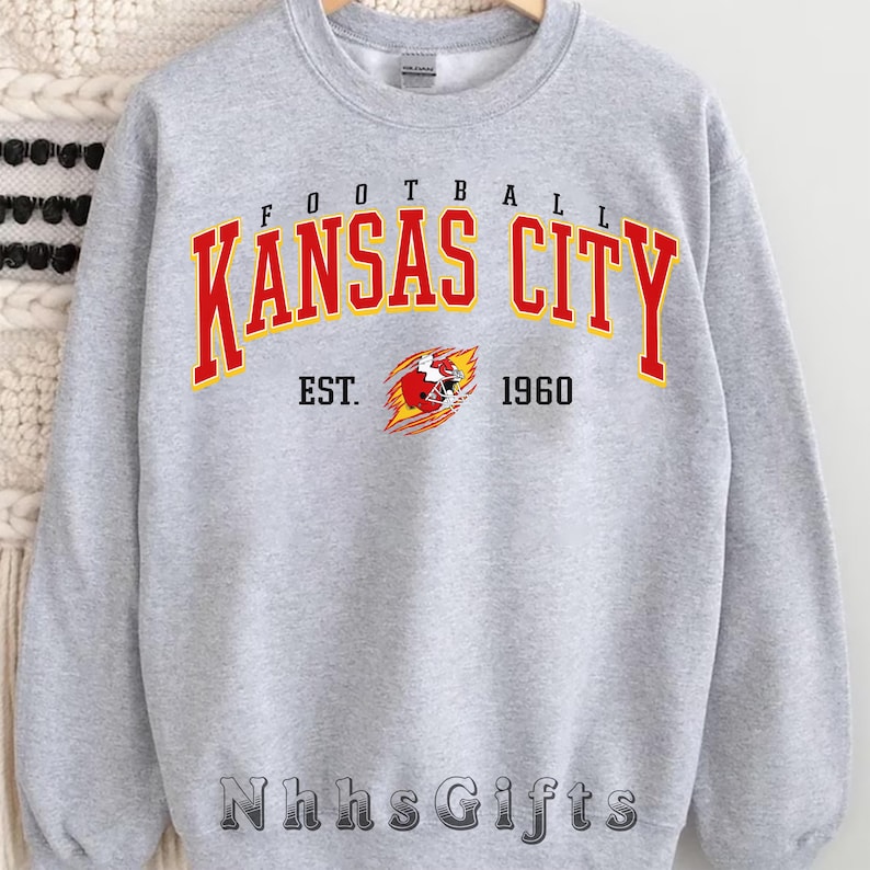 May include: A light gray sweatshirt with the words "FOOTBALL KANSAS CITY" in red and yellow lettering. Below the text is "EST. 1960" and a graphic of a football helmet. The brand name "Nhhs Gifts" is at the bottom.