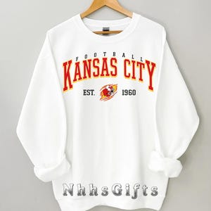 May include: A white sweatshirt with the words "FOOTBALL KANSAS CITY" in red and yellow lettering. Below the text is "EST. 1960" and a graphic of a football helmet. The sweatshirt is hanging on a wooden hanger.