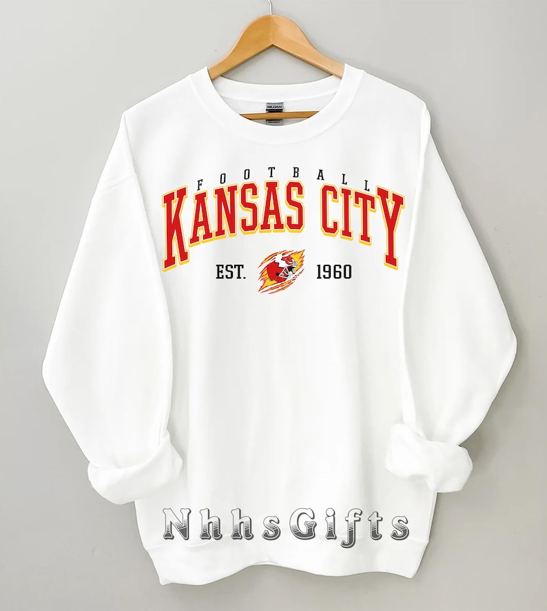 May include: A white sweatshirt with the words "FOOTBALL KANSAS CITY" in red and yellow lettering. Below the text is "EST. 1960" and a graphic of a football helmet. The sweatshirt is hanging on a wooden hanger.