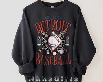 May include: Dark grey sweatshirt with "DETROIT BASEBALL" in orange and blue lettering, featuring a baseball graphic. The text "SINCE 1894" is above the word "DETROIT". The sweatshirt has rolled-up sleeves and the text "NhhsGifts" at the bottom.