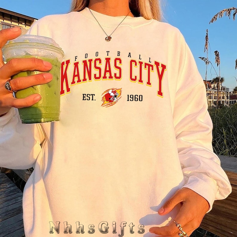 May include: White long-sleeve sweatshirt featuring "Kansas City" in red arched text with a yellow outline. Below the text is "EST. 1960" and a team logo. The sweatshirt has a crew neck and is a relaxed fit. The text "Nhhs Gifts" is at the bottom.