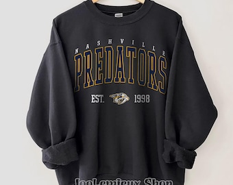 May include: Black crewneck sweatshirt with "NASHVILLE PREDATORS" in navy and gold lettering. The sweatshirt includes the team logo and "EST. 1998". The Gildan black sweatshirt is displayed on a wooden hanger.