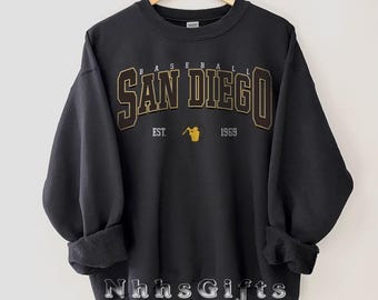 May include: Black sweatshirt featuring the text "SAN DIEGO BASEBALL" in gold and brown lettering. Below the text are the words "EST. 1969" with a gold baseball player graphic. The sweatshirt is displayed on a wooden hanger.