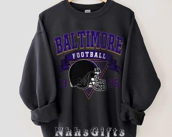 May include: A dark grey sweatshirt featuring the text "BALTIMORE FOOTBALL" in purple and gold, above a black football helmet graphic. The numbers "19" and "96" are on either side of the helmet. The text "NhhsGifts" is at the bottom.