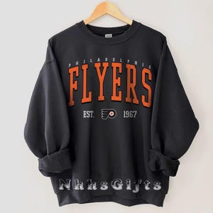 Può includere: Felpa nera con la scritta "PHILADELPHIA FLYERS" in lettere arancioni. Sotto, si legge "EST. 1967" con il logo della squadra. La felpa è appesa a una gruccia di legno.