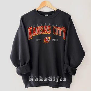 May include: Black sweatshirt with the words "KANSAS CITY" in red and gold lettering. The text "FOOTBALL" is above the city name, and "EST. 1960" is below, along with a team logo. The sweatshirt is hanging on a wooden hanger.