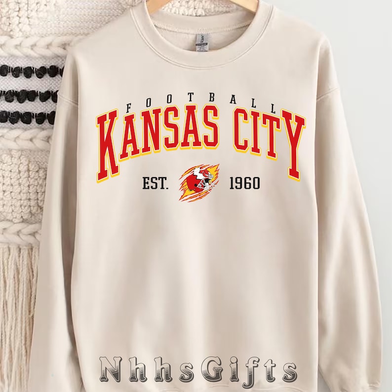 May include: Cream-colored sweatshirt featuring the text "Kansas City" in red and yellow, with "Football" above and "EST. 1960" below. A graphic of a football player is in the center. The brand name "Nhhs Gifts" is at the bottom.