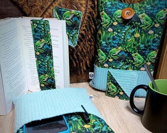 May include: A book cover with a green and black frog pattern. The cover has a blue pocket on the front and a brown button. A phone and pen are in the pocket. The cover is on a wooden table.
