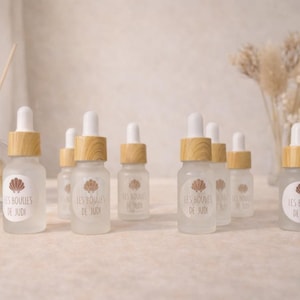 Peut inclure: Plusieurs petits flacons en verre dépoli avec des compte-gouttes blancs et des bouchons en bois. Chaque flacon porte une étiquette avec un motif de coquillage et le texte "LES BOUCLES DE JUDI". Un diffuseur et des fleurs séchées sont en arrière-plan.