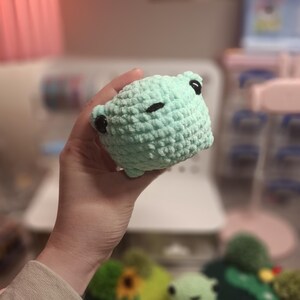 Puede incluir: Un peluche de rana de color verde claro tejido a crochet con ojos negros. La rana está siendo sostenida en una mano.