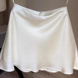 Puede incluir: Minifalda de satén color crema con una ligera forma de línea A. La falda tiene una textura suave y fluida y un brillo sutil, lo que sugiere una tela ligera. La falda está suspendida de una fina correa.
