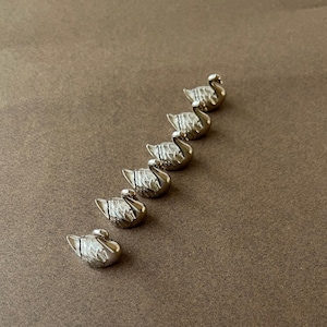 May include: Six small, silver-colored swan figurines arranged diagonally on a brown surface. Each swan has detailed feathering and a curved neck, giving them a graceful appearance. The figurines are likely decorative or part of a jewelry set.
