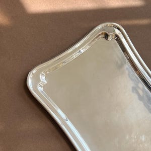 以下が含まれることがあります： 光沢のある反射面を持つ長方形の銀色のサービングトレイ。滑らかで盛り上がった縁と丸みを帯びた角が特徴です。トレイは清潔感のある金属仕上げで、光を反射するシンプルなデザインです。