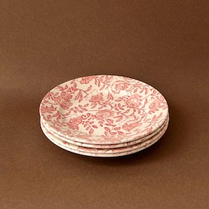 May include: Four white ceramic plates with a pink floral design are stacked. The plates display a repeating pattern of flowers and leaves. The plates are stacked on a brown background. Each plate measures approximately 20 cm in diameter.