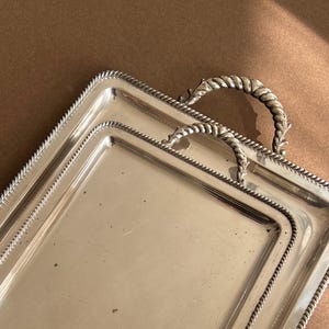 Peut inclure: Un plateau de service rectangulaire argenté avec une bordure en forme de corde et une poignée. Le plateau a une surface brillante et réfléchissante, probablement en métal. La poignée est argentée et présente un motif torsadé.