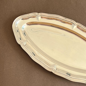 Può includere: Un vassoio da portata ovale color argento con una superficie lucida e riflettente e un bordo rialzato smerlato. Il vassoio presenta una base centrale liscia e allungata e un design del bordo curvo che segue la forma ovale del piatto.