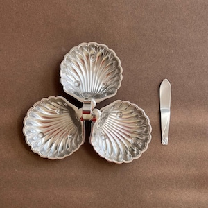 May include: A silver-toned metal serving dish featuring three connected shell-shaped bowls arranged in a clover pattern, each with a ribbed interior and three small feet. A matching flat silver-toned spreader knife is positioned to the right.