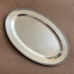 Puede incluir: Una bandeja de servicio ovalada de tono plateado con una superficie central lisa y reflectante, y un borde decorativo con cuentas a lo largo del contorno exterior. La bandeja de metal presenta un acabado pulido y un diseño sencillo.