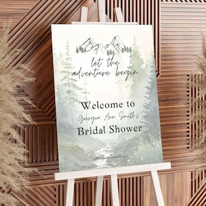 May include: A white easel displays a sign with a watercolor forest scene and mountains. The text reads "let the adventure begin" and "Welcome to Georgia Ann Smith's Bridal Shower". The sign has a hashtag: #lettheadventurebegin.