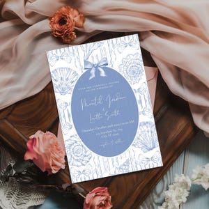 May include: Wedding invitation with a blue oval containing names and event details. The invitation features a white background with blue seashell and bow illustrations. The invitation is surrounded by flowers and fabric.