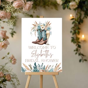Può includere: Un cartello di benvenuto per un addio al nubilato su un cavalletto. Il cartello presenta illustrazioni di stivali da cowboy, fiori e un calice di champagne. Il testo recita "Benvenuti all'addio al nubilato di Elizabeth."