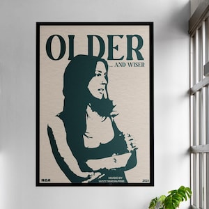 Peut inclure: Une affiche encadrée avec les mots "OLDER...AND WISER" en gras, en vert foncé. L'affiche présente une silhouette verte d'une femme. L'affiche est accrochée à un mur blanc au-dessus d'une table en bois et de chaises noires.