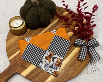 May include: A Halloween-themed dog bandana with orange, black and white patterns, and the text "BOO". The bandana is displayed on a wooden cutting board with a green pumpkin, a candle, and red decorative branches.