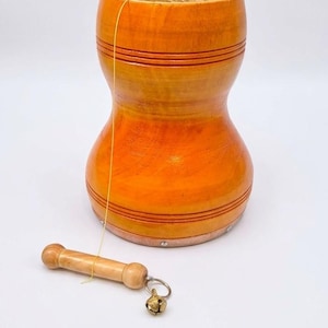Può includere: Un tamburo in legno marrone chiaro lucidato, a forma di clessidra con linee orizzontali scanalate. Un sottile filo giallo collega il tamburo a una piccola impugnatura in legno con un campanello metallico fissato a un anello argentato.