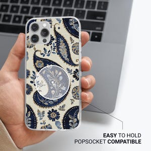 May include: Clear phone case featuring a navy blue and light blue paisley pattern on a beige backdrop. The case is labelled "EASY TO HOLD POPSOCKET COMPATIBLE". The phone case is held in a person's hand.