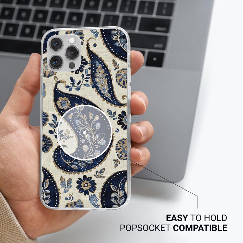May include: Clear phone case featuring a navy blue and light blue paisley pattern on a beige backdrop. The case is labelled "EASY TO HOLD POPSOCKET COMPATIBLE". The phone case is held in a person's hand.