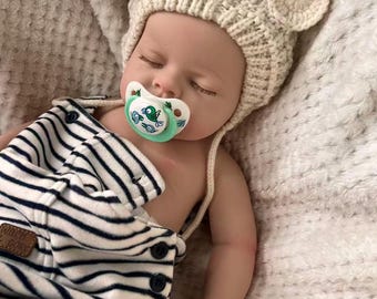 Puede incluir: Un bebé dormido con un gorro de punto color crema con orejas de oso y un pelele a rayas azul marino y blanco. El bebé tiene un chupete en la boca. El pelele tiene un pequeño parche marrón con texto.