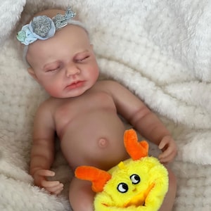 May include: A lifelike baby doll with closed eyes, wearing a floral headband. The doll is nude, with a yellow plush toy with orange accents covering its lower body. The doll is lying on a white, fluffy blanket.