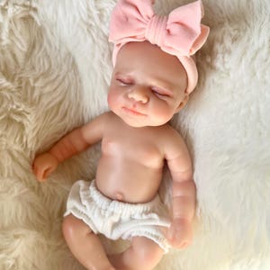 Op de afbeelding: Een levensechte babypop met gesloten ogen, met een roze strik-hoofdband en een witte luier. De pop ligt op een zacht, wit oppervlak van imitatiebont. De huid van de pop heeft een realistische tint.