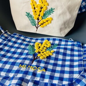 Peut inclure: Trousse de maquillage crème et tablier vichy bleu et blanc, tous deux brodés de fleurs de mimosa jaunes et de feuilles vertes. Le mot "MIMOSA" est brodé en jaune sur les deux articles.