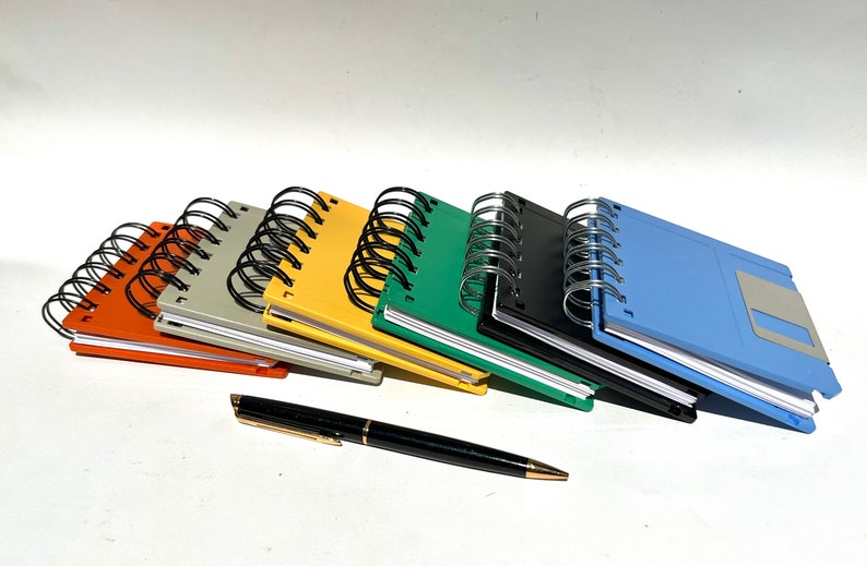 May include: A set of seven colorful spiral bound notebooks, each with a different color cover. The notebooks are stacked on top of each other, with the top notebook being blue. The notebooks are all the same size and have a vintage floppy disk design. A black pen is lying on the table in front of the notebooks.