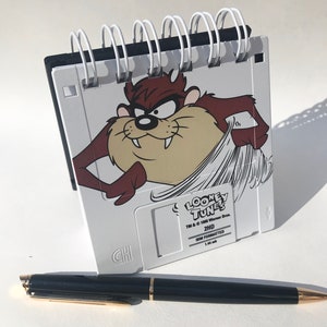 May include: A white floppy disk with a black and white spiral binding. The disk has a cartoon image of Taz from Looney Tunes on the front. The disk is labeled "Looney Tunes 2HD IBM Formatted 1.44 Mb".