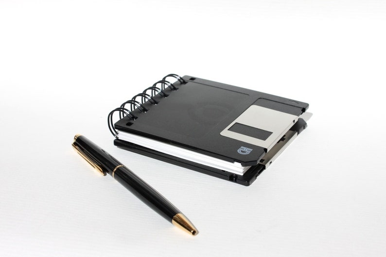 May include: A black spiral notebook with a black floppy disk on top of it. A black pen with a gold tip is lying next to the notebook.