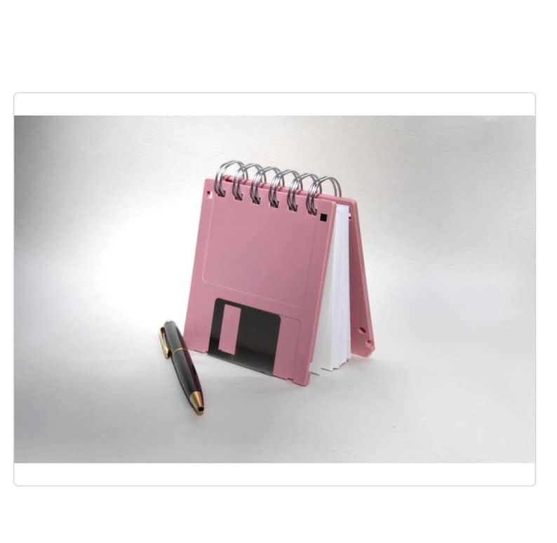 May include: A pink floppy disk shaped notebook with a silver spiral binding. The notebook is closed and sitting on a white surface with a black pen next to it.