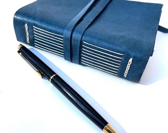 Può includere: Un diario in pelle blu navy con chiusura a cinturino abbinata e cuciture bianche. Una penna nera con dettagli dorati è appoggiata accanto al diario. La penna ha la punta dorata e una fascia dorata.