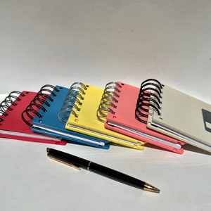 May include: Five colorful spiral bound notebooks with a black pen. The notebooks are stacked on top of each other and have a vintage floppy disk design.