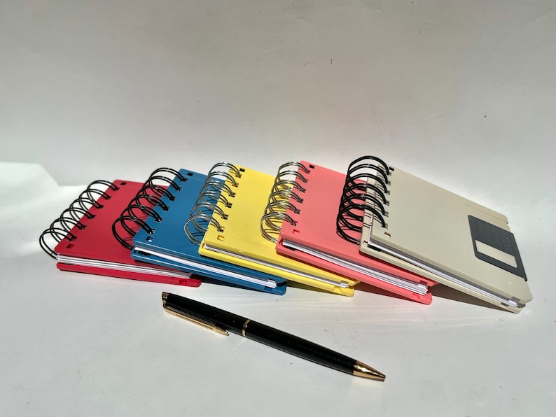 May include: Five colorful spiral bound notebooks with a black pen. The notebooks are stacked on top of each other and have a vintage floppy disk design.