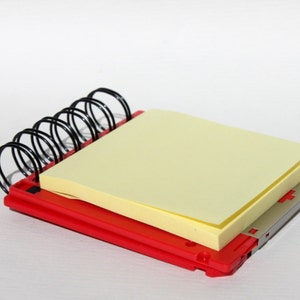 May include: A red spiral notebook with a yellow sticky note on top. The notebook is made of plastic and has a silver metal binding.