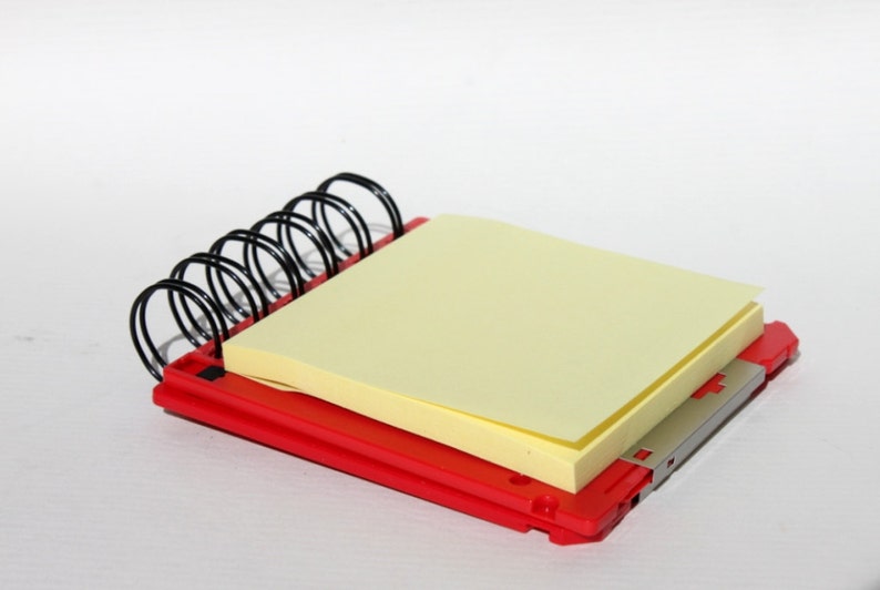 May include: A red spiral notebook with a yellow sticky note on top. The notebook is made of plastic and has a silver metal binding.