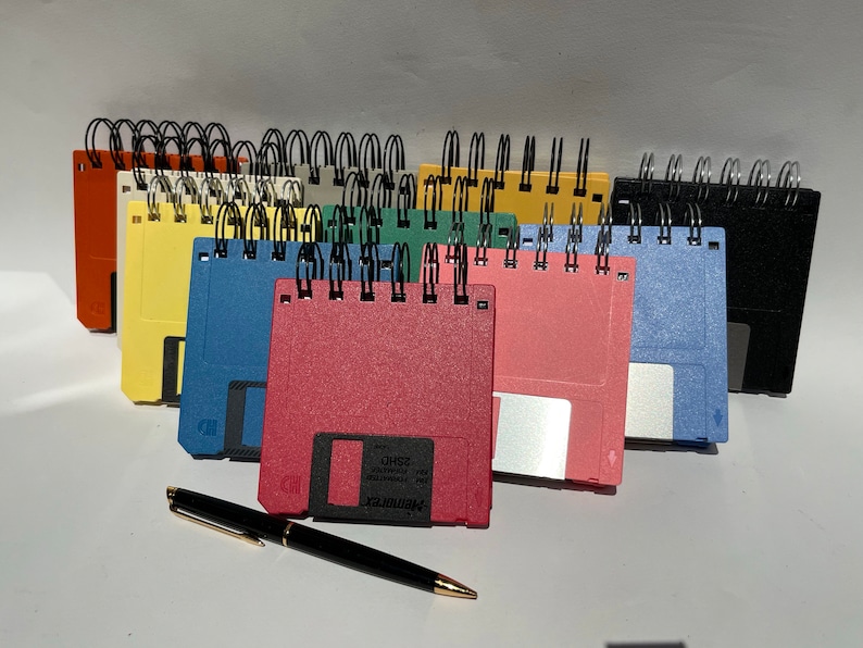 May include: A collection of colorful floppy disk notebooks with black metal spiral binding. The notebooks are arranged in a row, with the covers facing the viewer. The notebooks are all different colors, including red, pink, blue, green, yellow, and orange. A black pen is lying on the table in front of the notebooks.